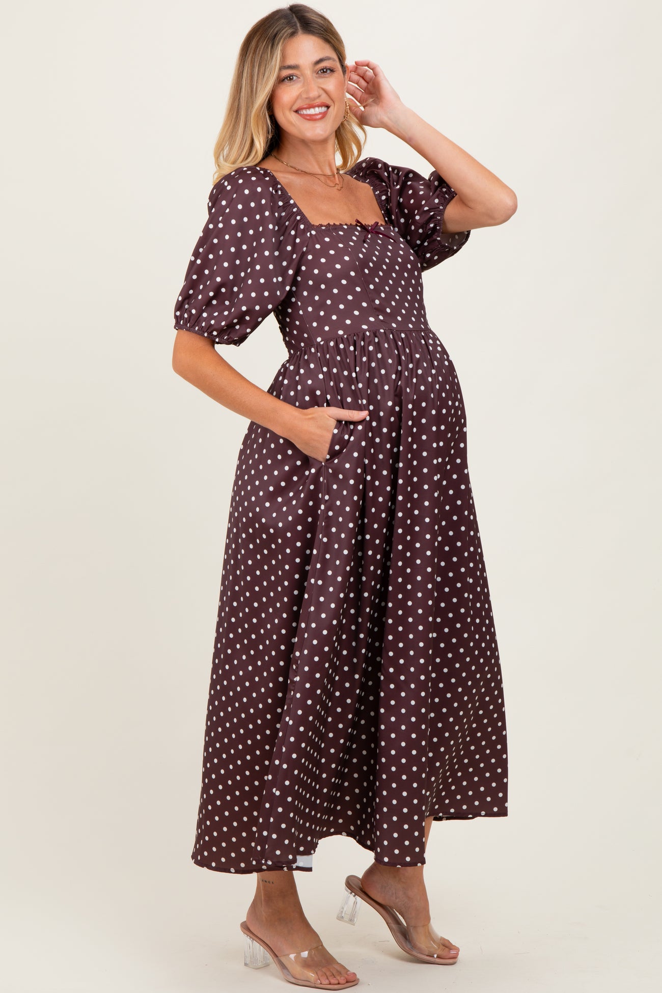 woman wearing a brown polka dot maternity dress with puff sleeves and pockets, showcasing her baby bump