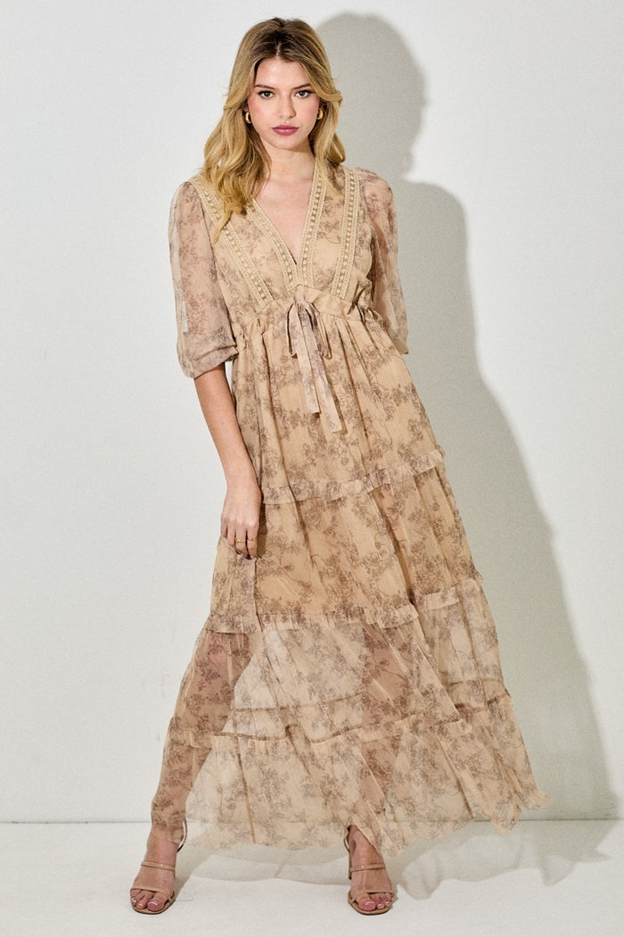 woman wearing a beige floral maxi dress with sheer layers and a tie waist, standing against a white background