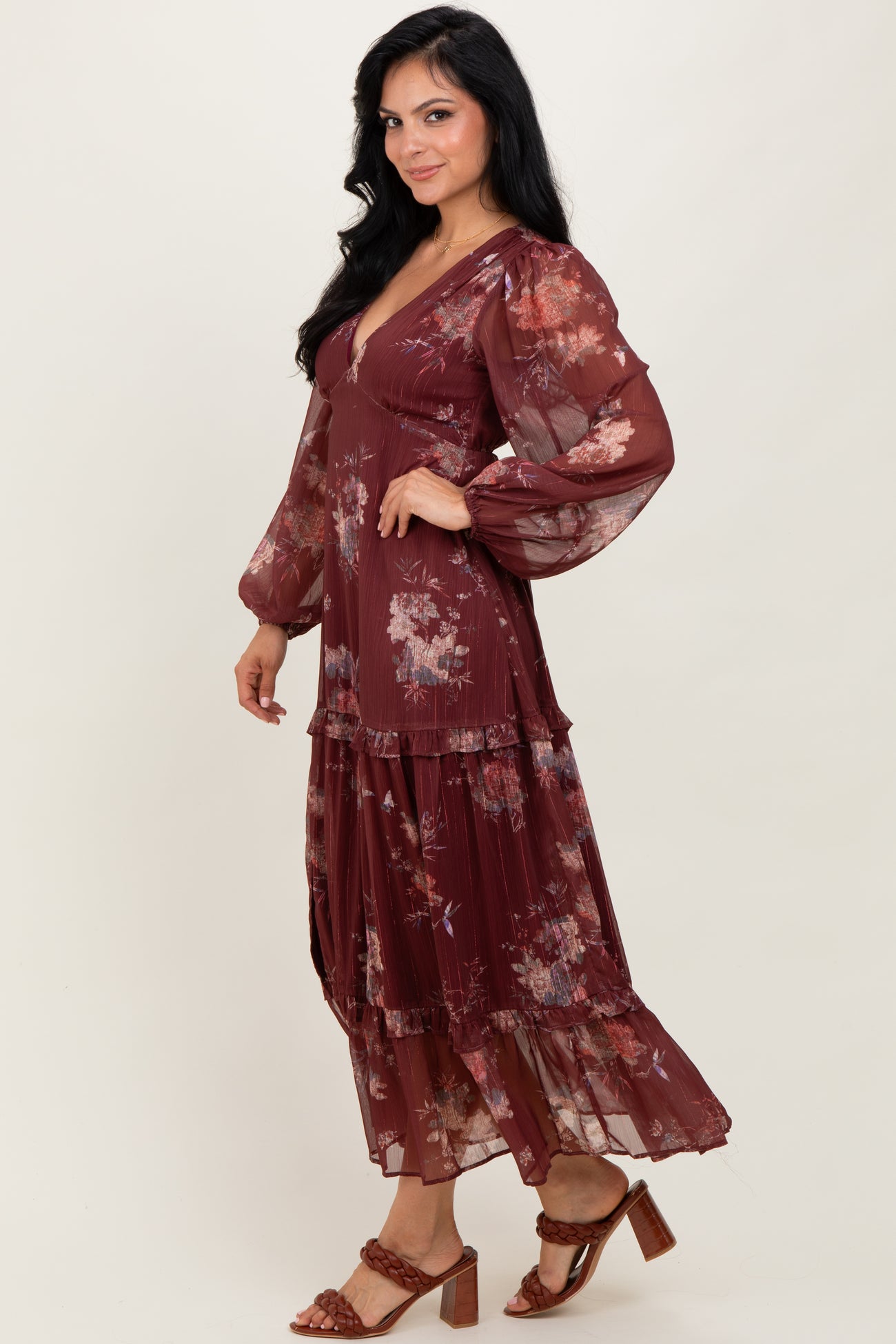 woman wearing a burgundy floral maxi dress with long sleeves and tiered ruffles, standing sideways against a neutral background