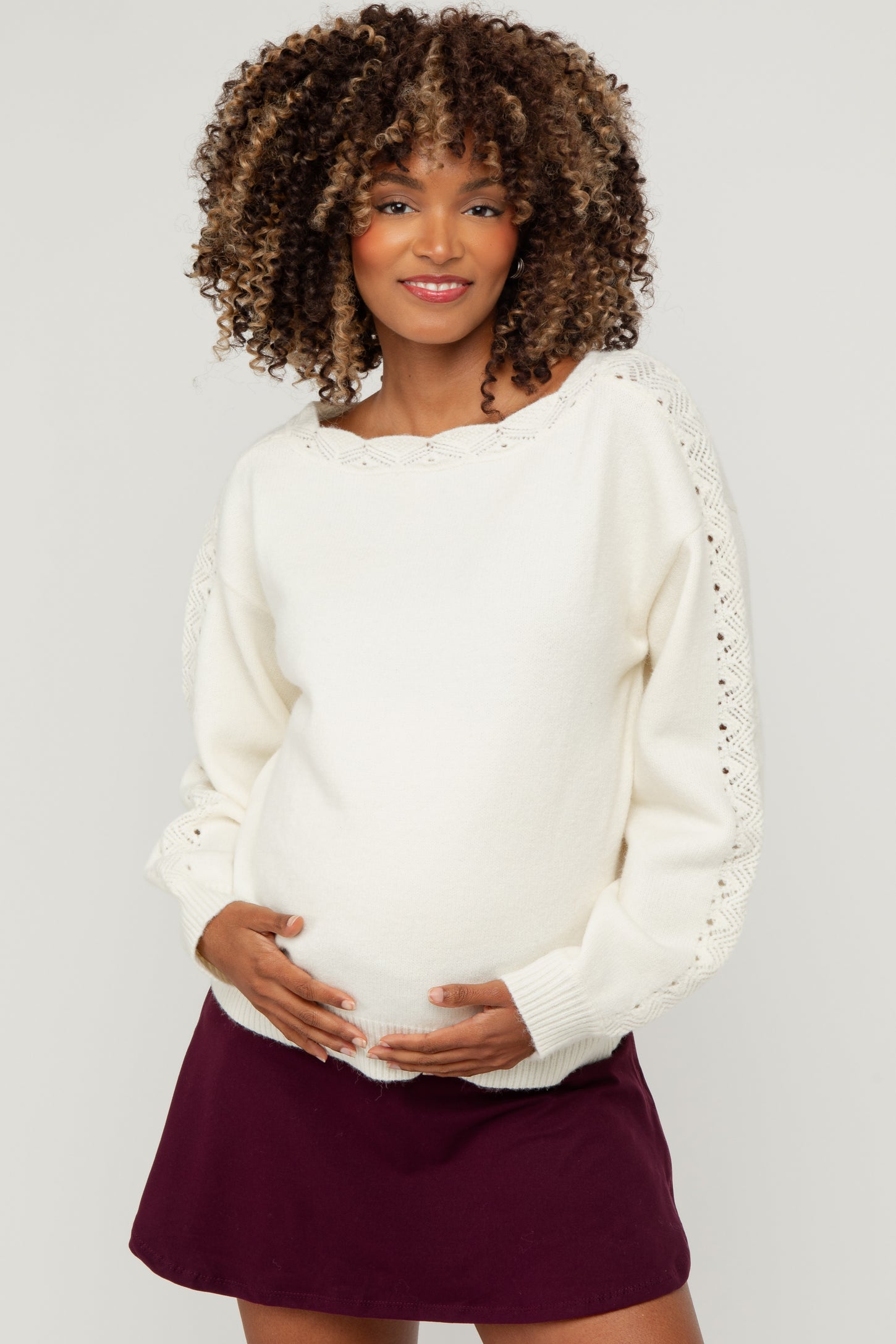woman wearing a cream maternity sweater with decorative sleeves and holding her baby bump