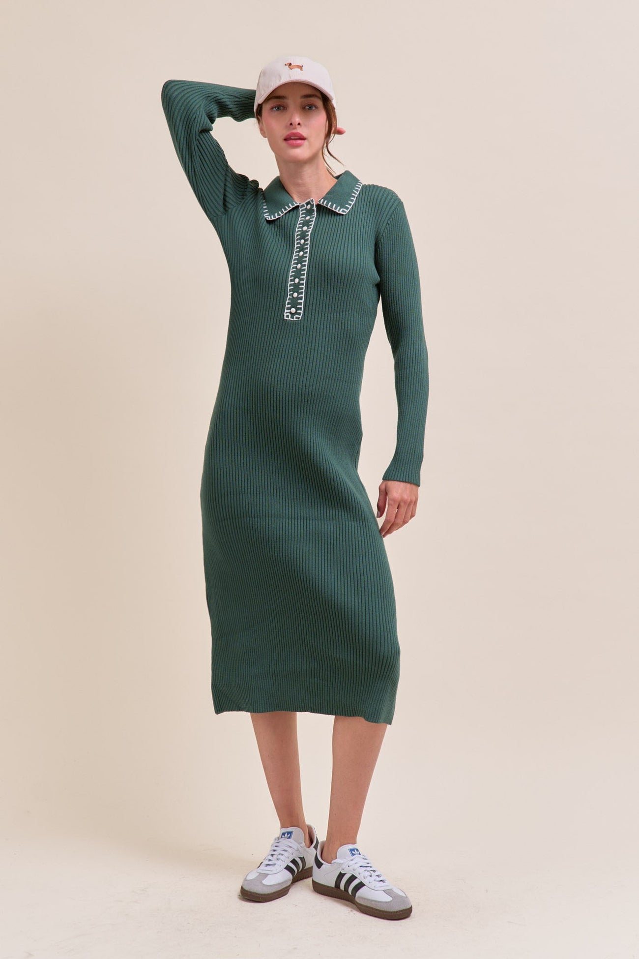 woman wearing a dark green ribbed midi dress with a collared neckline and long sleeves, styled with a baseball cap and sneakers