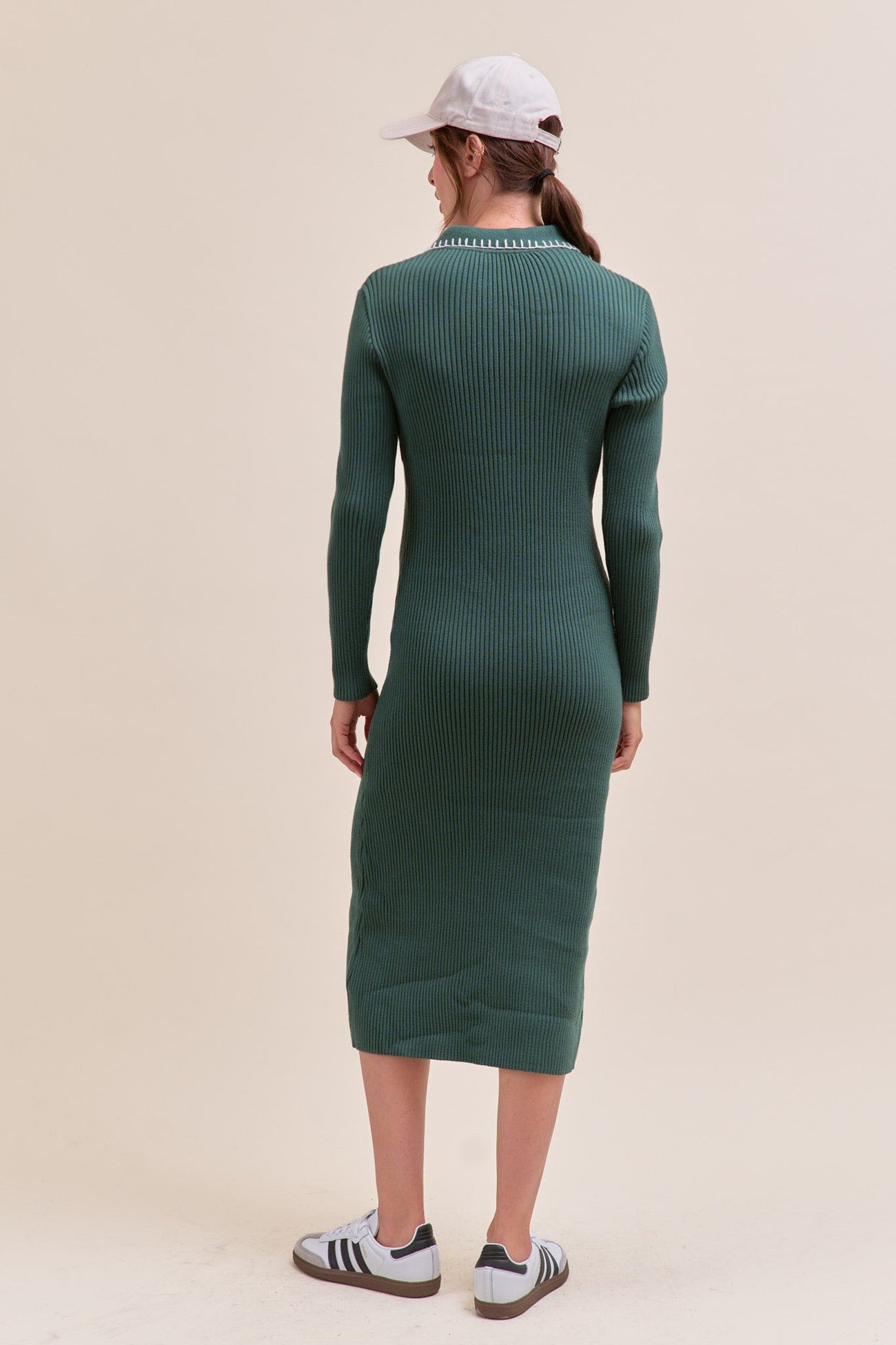 woman wearing a dark green ribbed midi dress with long sleeves, viewed from the back, paired with a white cap and sneakers
