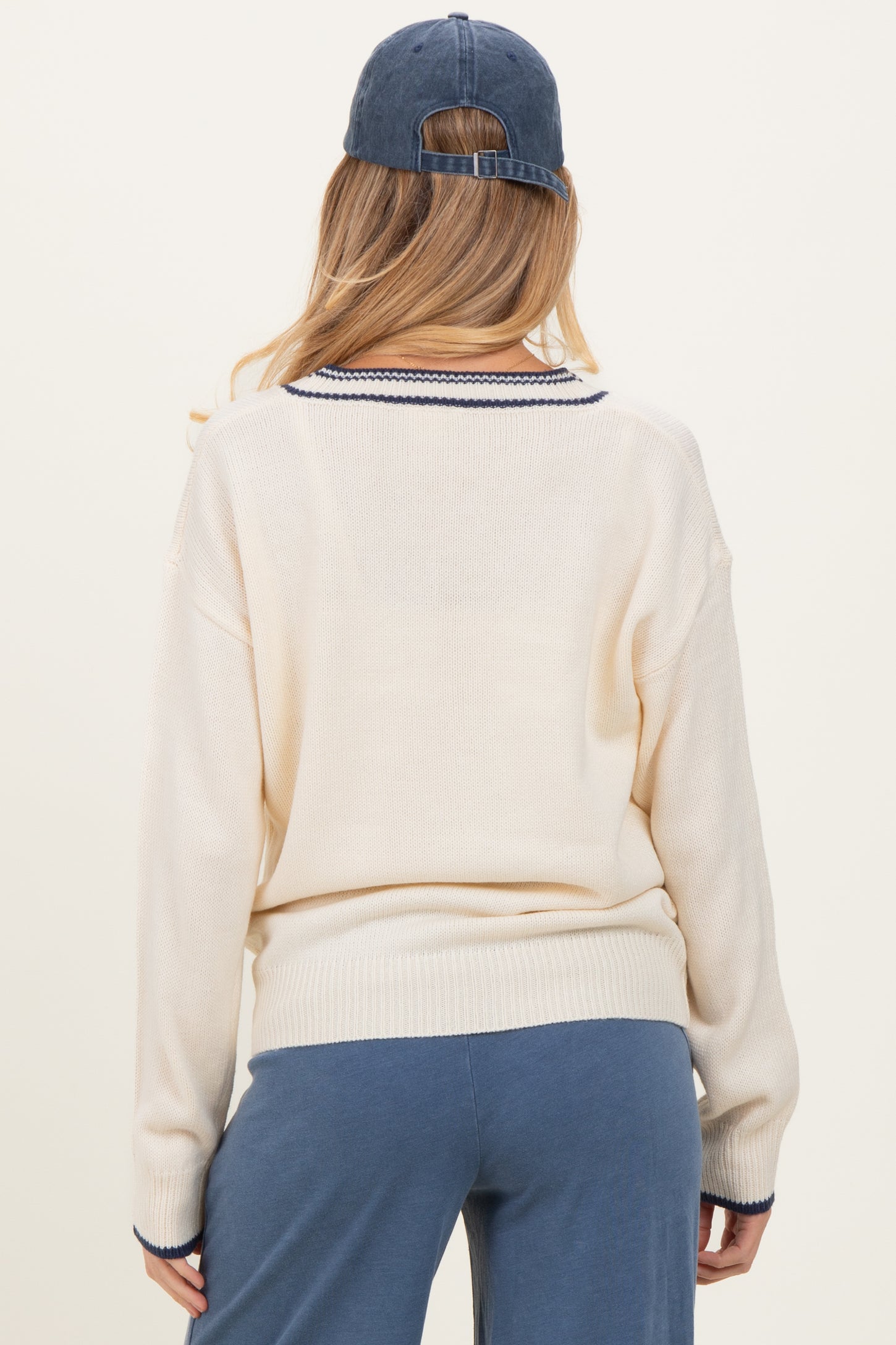 woman wearing a cream-colored maternity sweater with navy blue trim and back turned, paired with blue pants and a cap