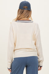 woman wearing a cream-colored maternity sweater with navy blue trim and back turned, paired with blue pants and a cap