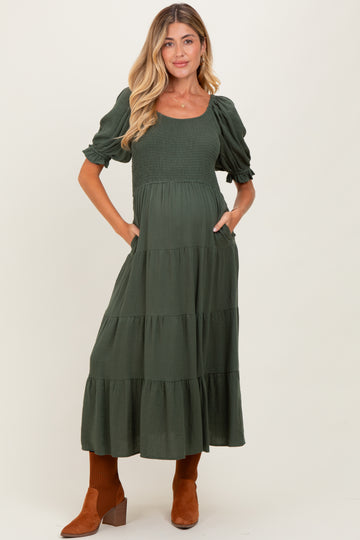 woman wearing a dark green maternity maxi dress with puff sleeves and tiered skirt, holding her baby bump