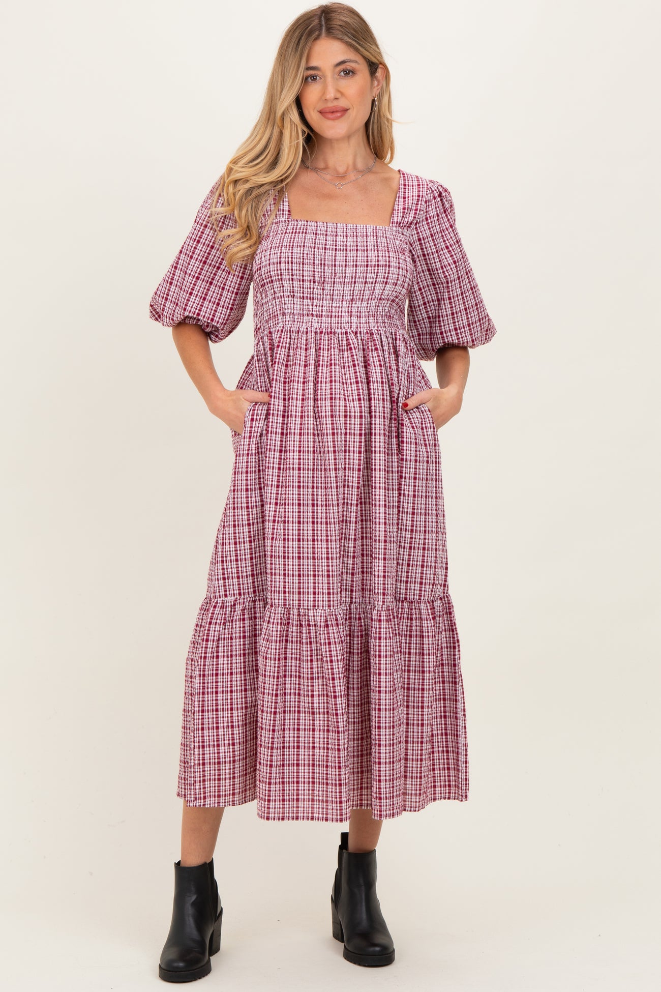 woman wearing a pink and white gingham maternity midi dress with puff sleeves and pockets, showcasing her baby bump