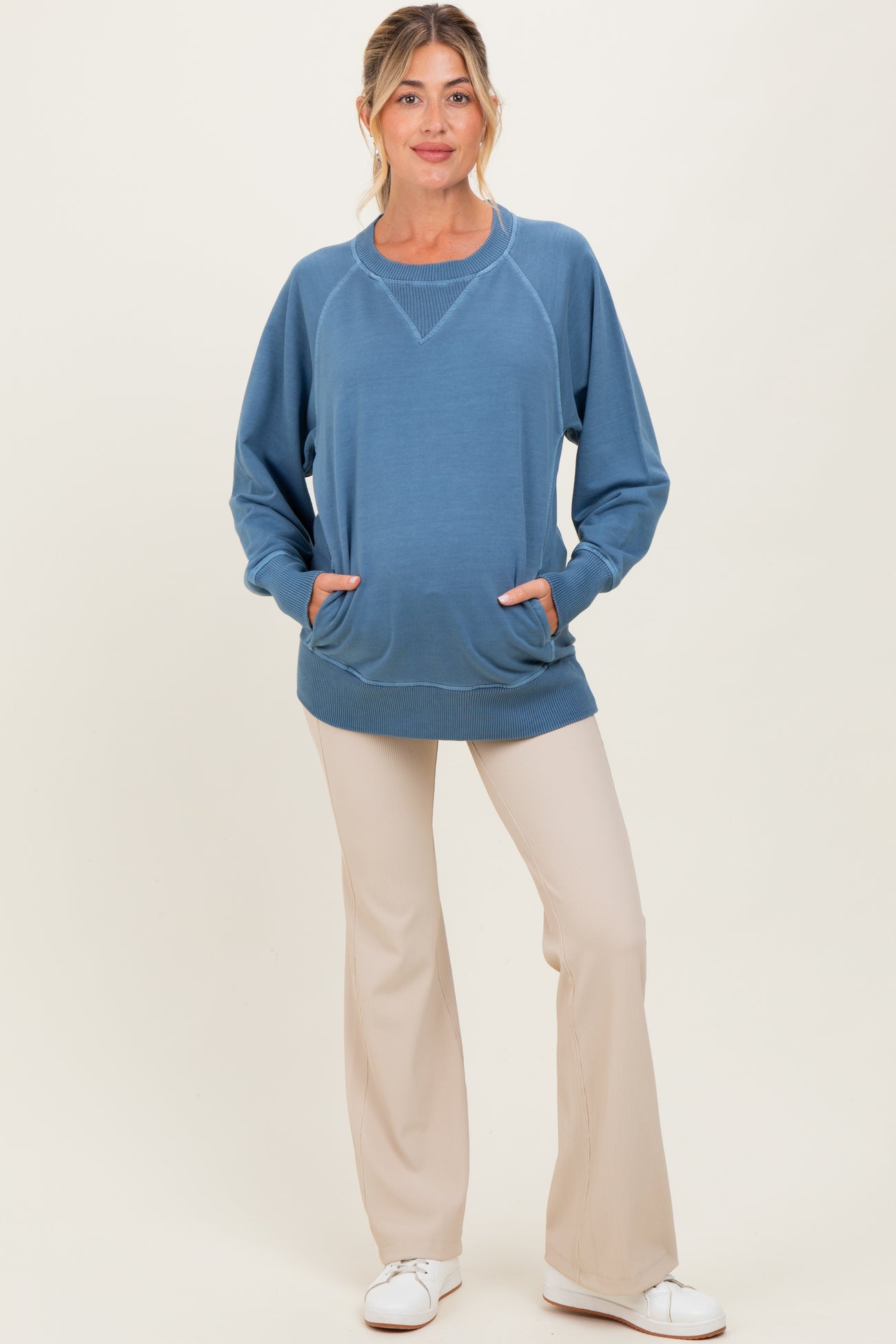woman wearing a blue maternity sweatshirt with a relaxed fit and front pockets, paired with light beige pants and a baby bump visible