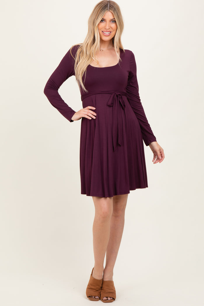 woman wearing a burgundy long sleeve dress with a tie waist and flared skirt, standing in a neutral background