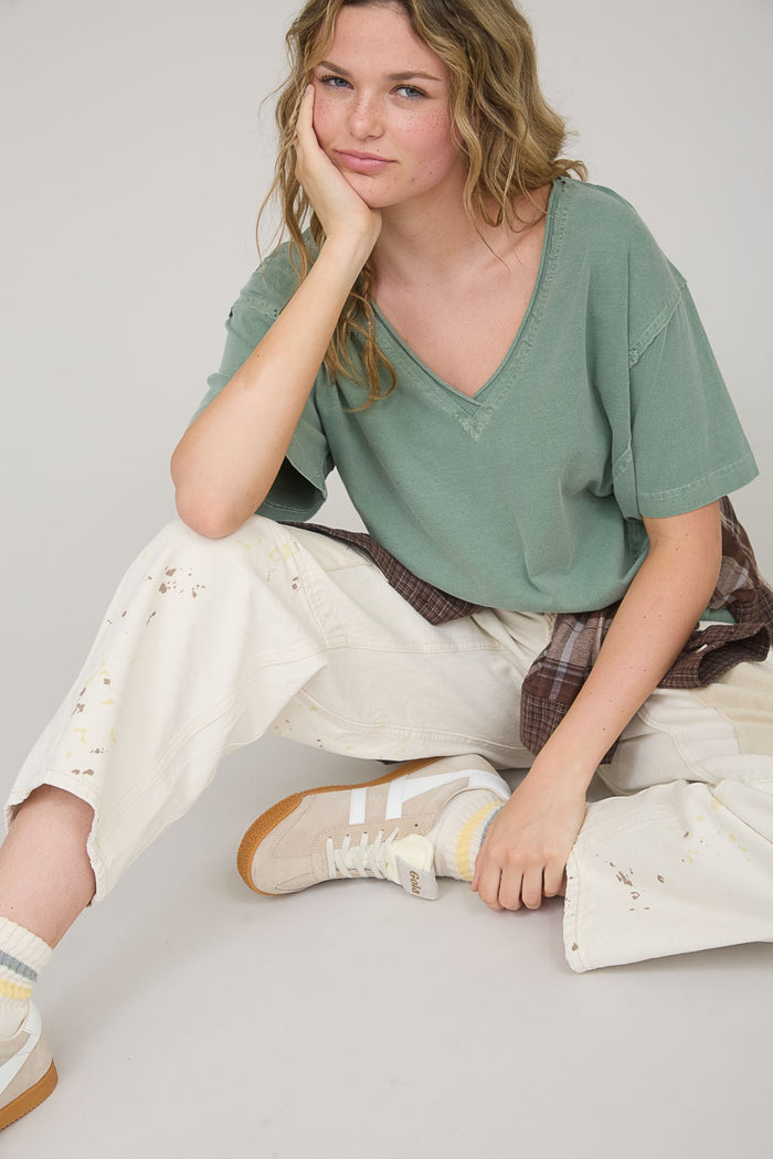 woman sitting in a relaxed pose wearing a green v-neck t-shirt, off-white distressed pants, and beige sneakers