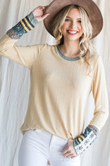 woman wearing a beige ribbed long sleeve top with patterned sleeves and a wide-brimmed hat, smiling at the camera