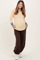 woman wearing a cream ribbed maternity top with decorative sleeves and brown maternity jogger pants, showing her baby bump