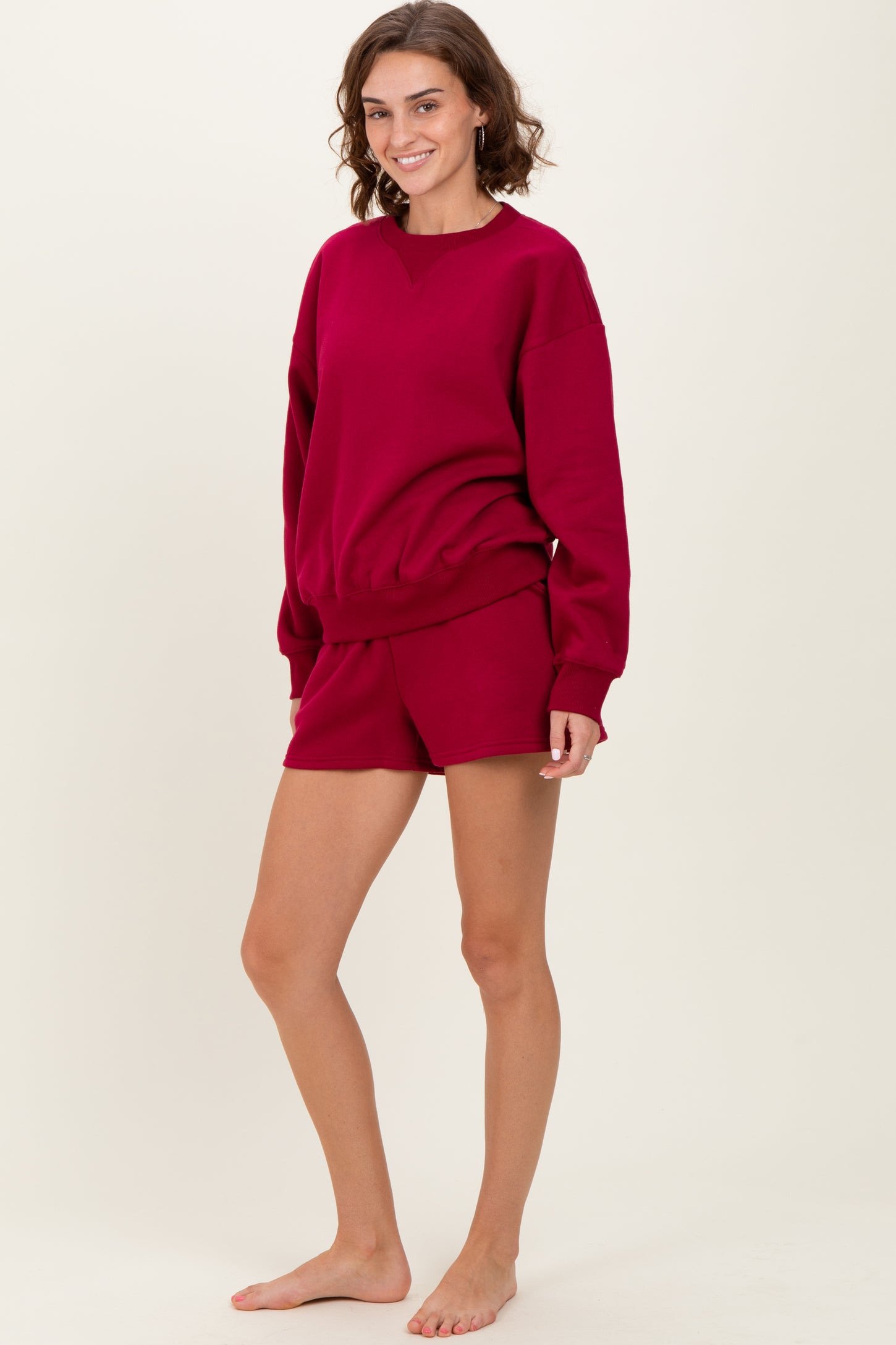 woman wearing a burgundy oversized sweatshirt and matching shorts, standing barefoot on a neutral background