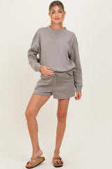 woman wearing a grey maternity sweatshirt and matching shorts, holding her baby bump while standing on a neutral background