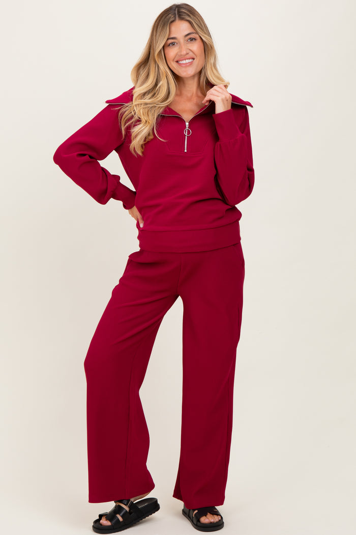 woman wearing a burgundy maternity athletic set with a zip-up sweatshirt and wide-leg pants, posing with one hand on her hip