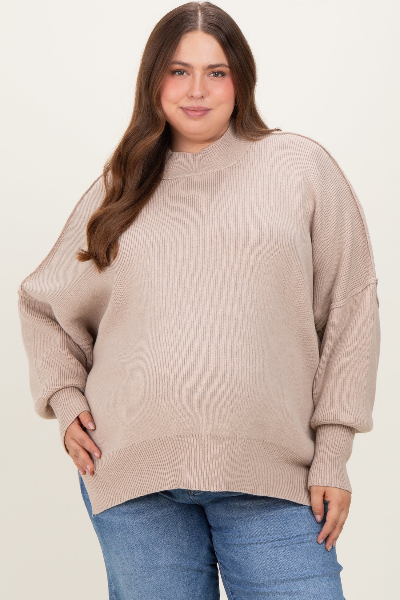 woman wearing a light beige oversized maternity plus sweater with a high neck and relaxed fit, styled with blue jeans