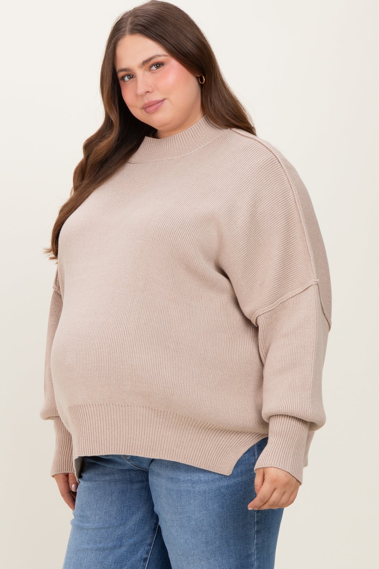 woman wearing a beige oversized maternity sweater with a high neckline and side slits, showcasing her baby bump