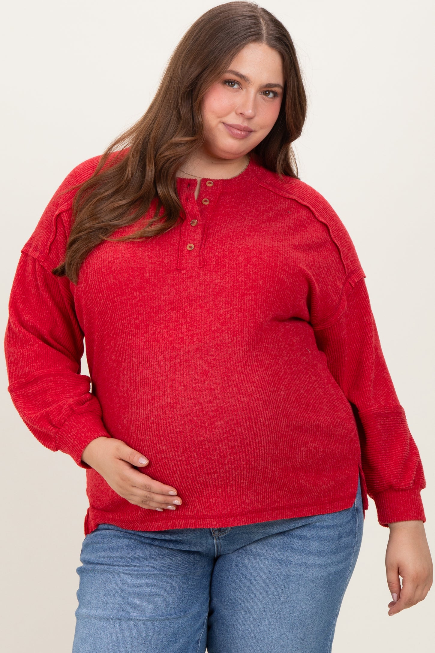 woman wearing a red long sleeve maternity top with buttons, holding her baby bump and paired with blue jeans