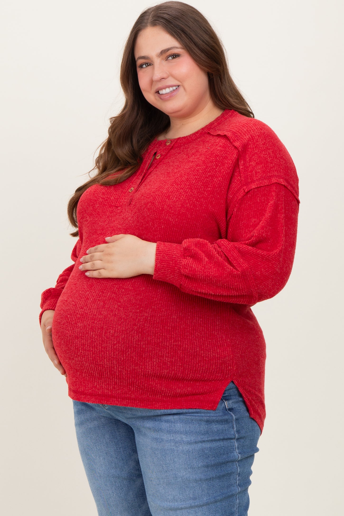 woman wearing a red maternity plus pullover with buttons, holding her baby bump and smiling