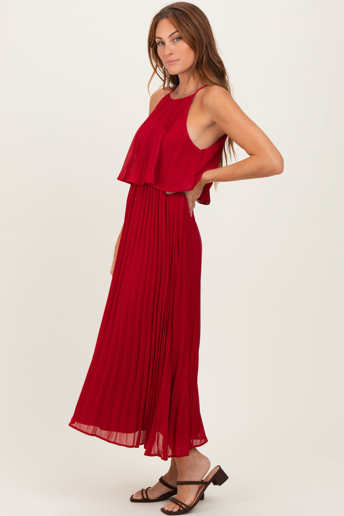 woman wearing a red pleated maxi dress with a layered design and thin straps, standing with one hand on her hip