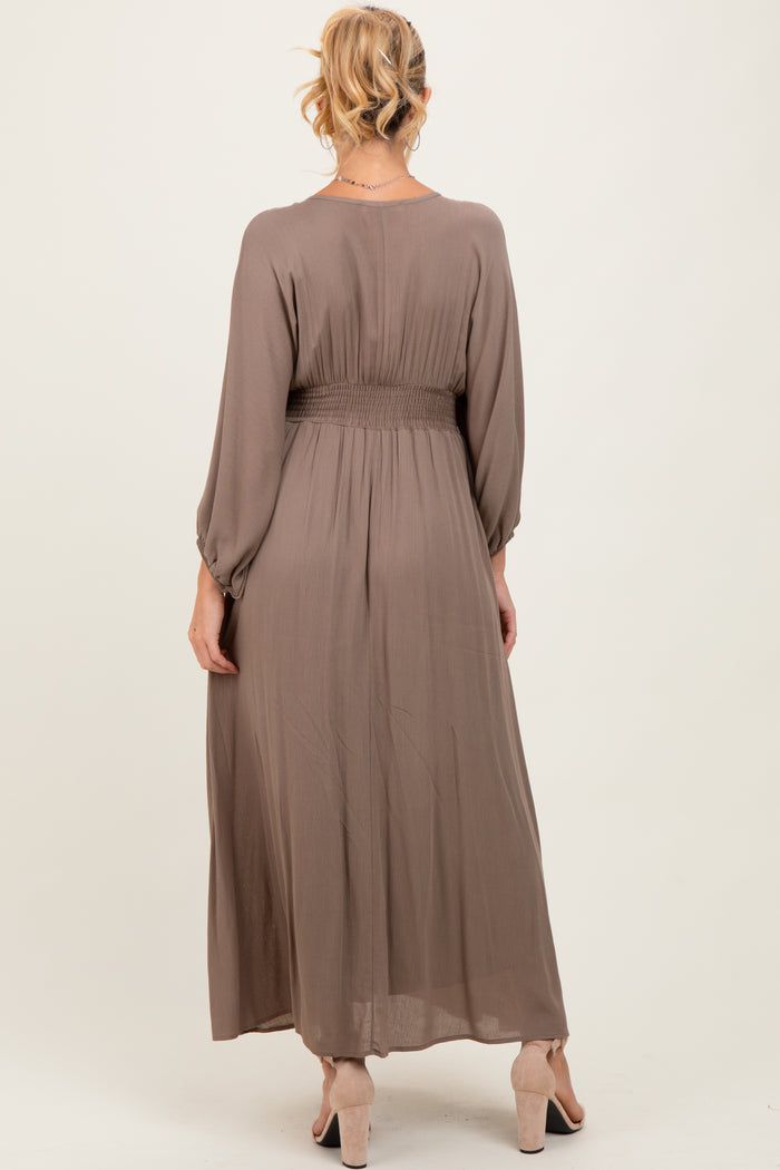 woman wearing a long taupe maternity dress with a smocked waist and long sleeves, viewed from the back