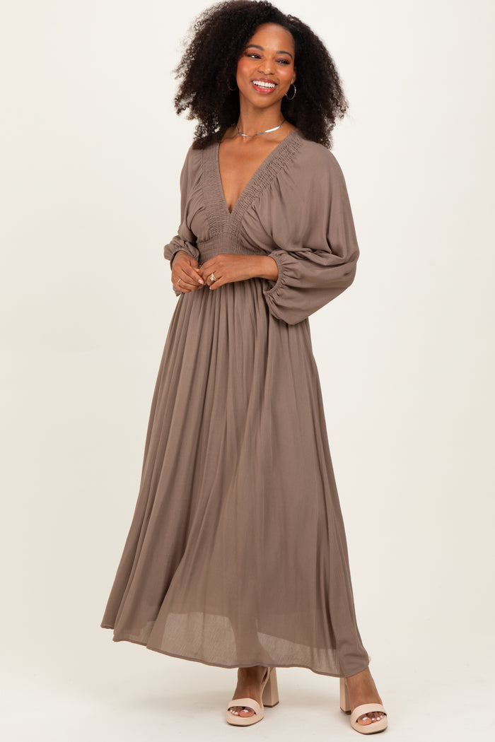 woman wearing a taupe long sleeve maxi dress with a deep v-neck and smocked bodice, standing and smiling
