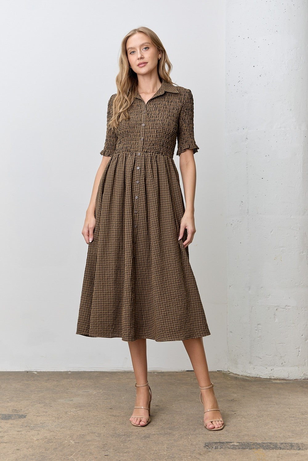 woman wearing a brown checkered midi dress with a smocked bodice and short sleeves, standing on a concrete floor