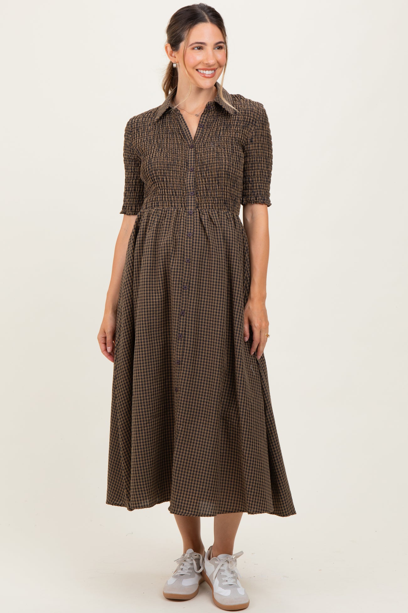 woman wearing a brown checked maternity dress with a button-down front and smocked bodice, holding her baby bump