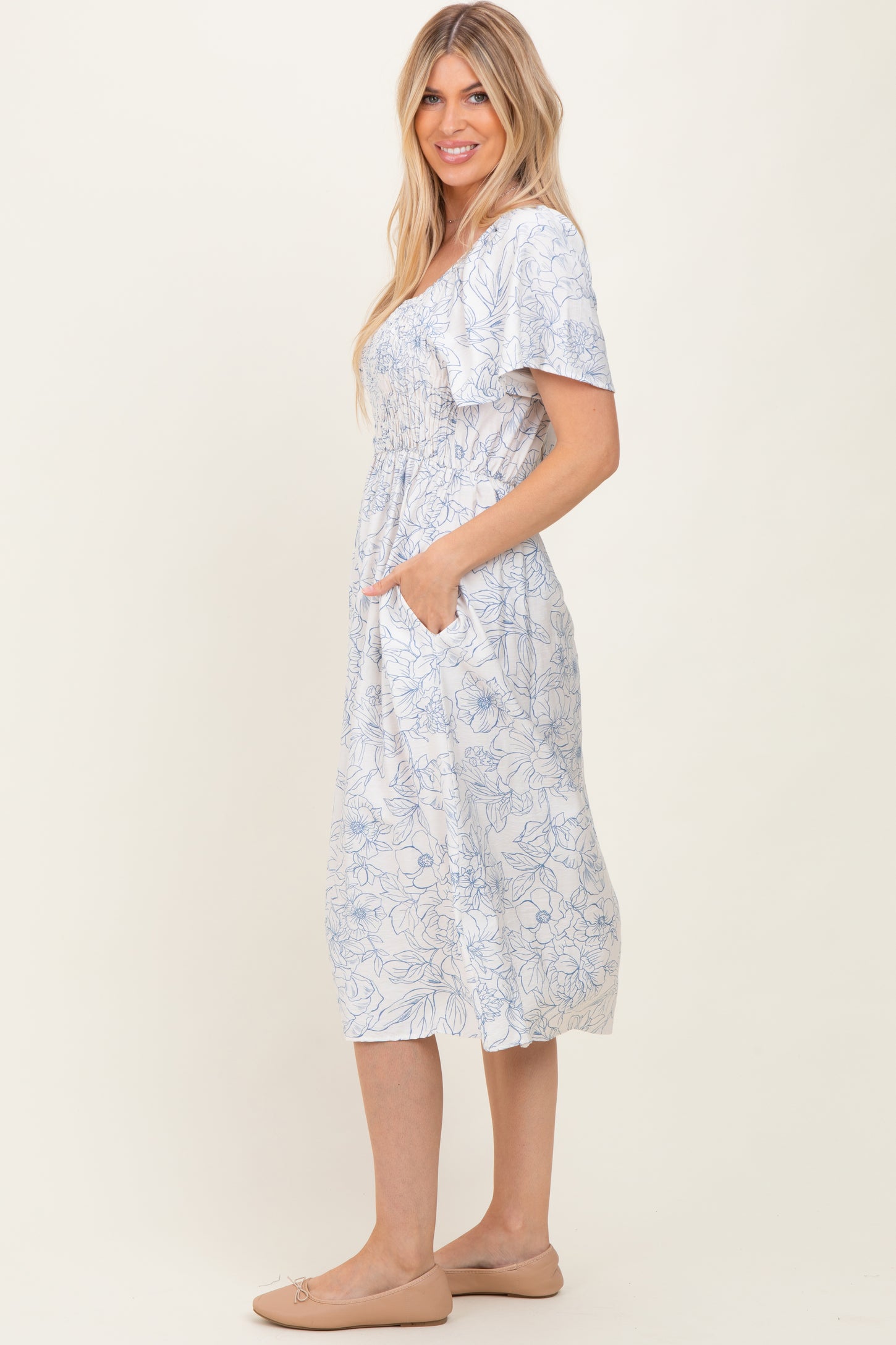 woman wearing a light blue floral midi dress with smocked bodice and short sleeves, standing with hands in pockets