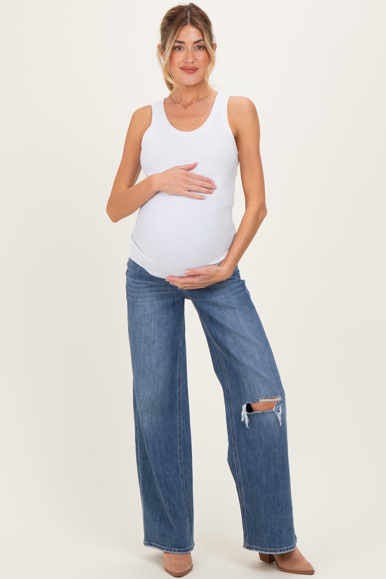 woman wearing a white ribbed maternity tank top and distressed wide-leg jeans, holding her baby bump