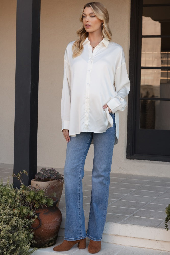 woman wearing a white satin maternity shirt and blue flared jeans, showcasing her baby bump while standing outdoors