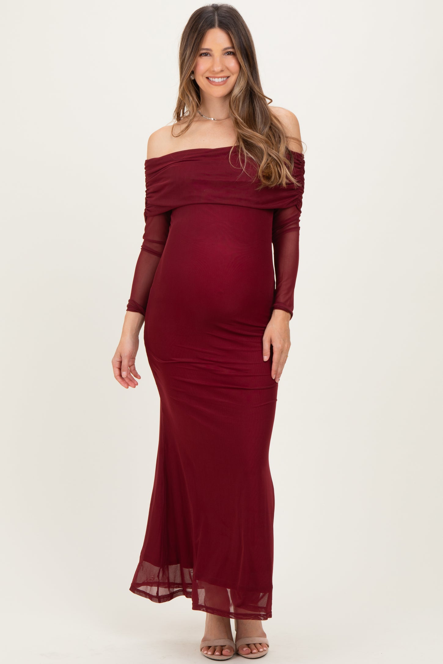 woman wearing a burgundy off-the-shoulder maternity gown with long sheer sleeves and a fitted silhouette, showcasing her baby bump