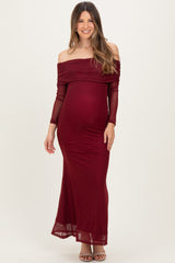 woman wearing a burgundy off-the-shoulder maternity gown with long sheer sleeves and a fitted silhouette, showcasing her baby bump