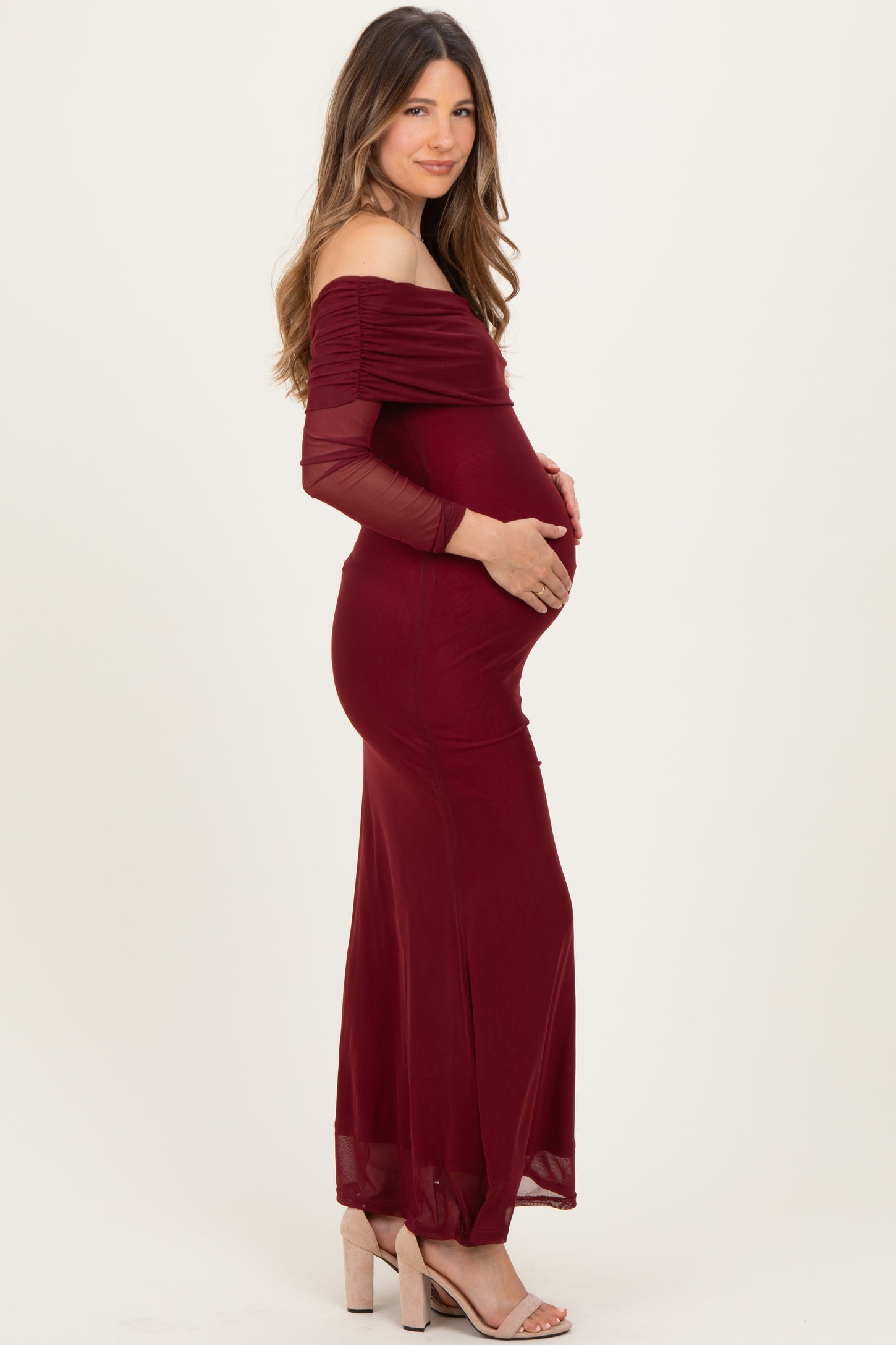 woman wearing a burgundy off-the-shoulder maternity gown with ruched sleeves and holding her baby bump