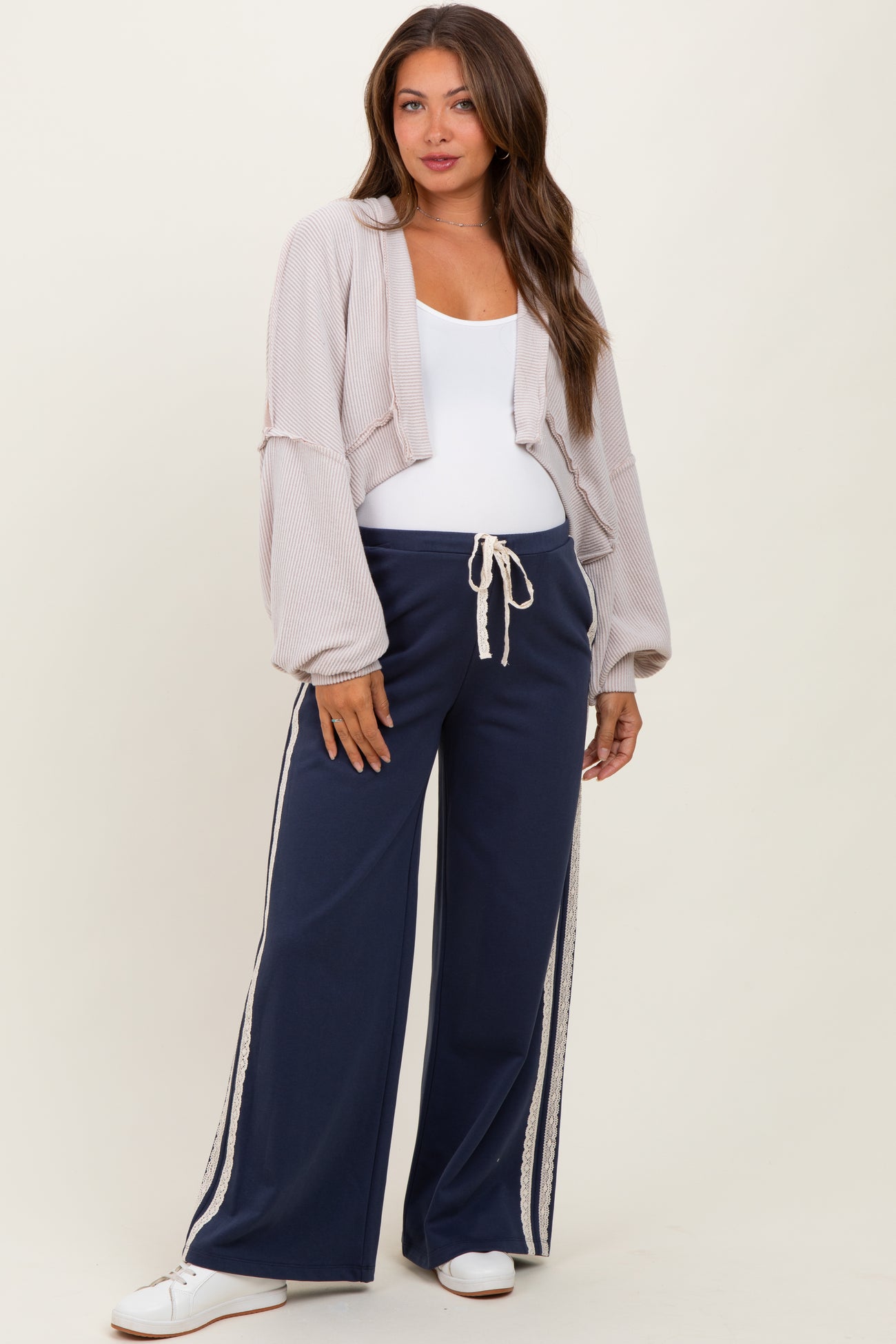 woman wearing a cropped light gray cardigan and navy maternity pants with white side stripes and drawstring