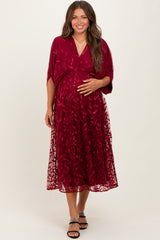 woman wearing a burgundy maternity dress with floral embroidery and a flowy overlay, holding her baby bump