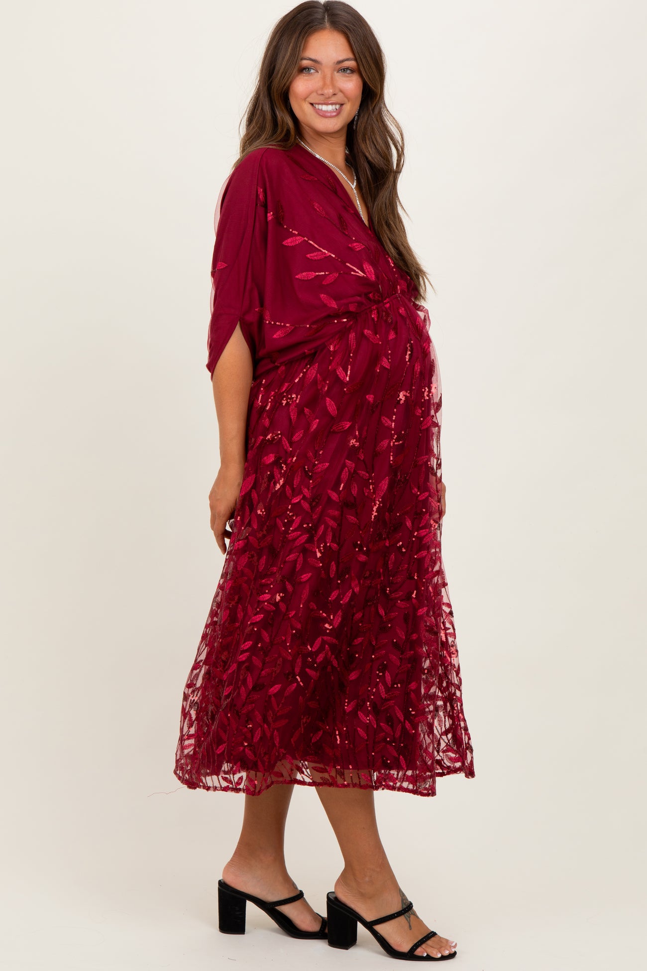 woman wearing a burgundy maternity dress with a floral lace overlay and a flowing silhouette, showcasing her baby bump