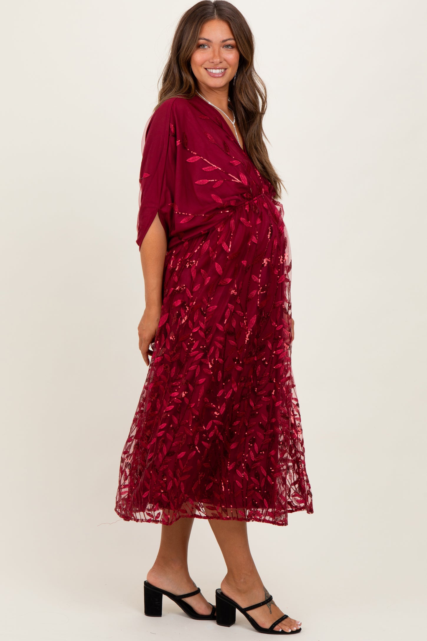 woman wearing a burgundy maternity dress with a floral lace overlay and a flowing silhouette, showcasing her baby bump