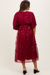 woman wearing a burgundy maternity dress with a floral sheer overlay and cinched waist, showcasing a pregnant belly