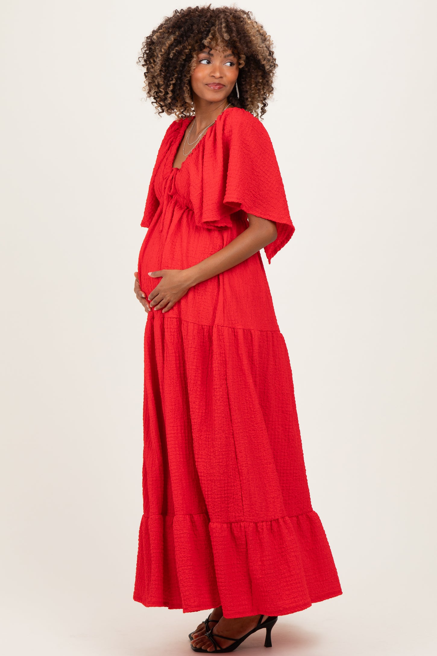 woman wearing a red tiered maternity dress with short sleeves and a bow tie, holding her baby bump