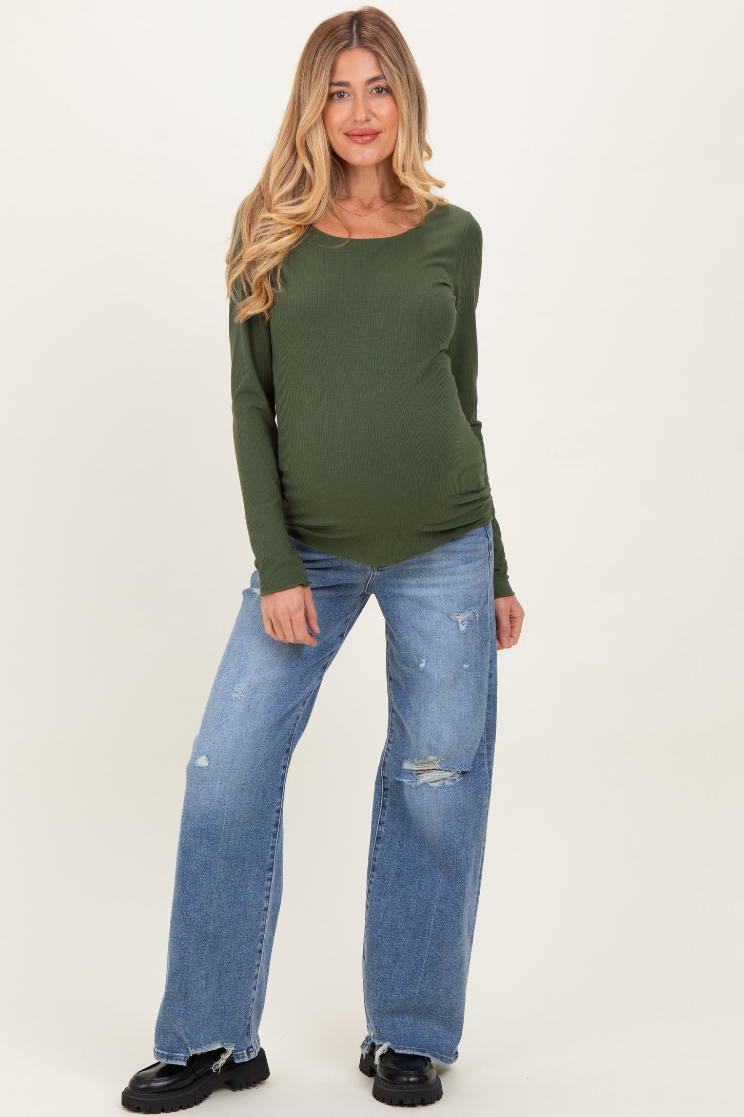 woman wearing a dark green long sleeve maternity top and distressed blue maternity jeans, showcasing her baby bump