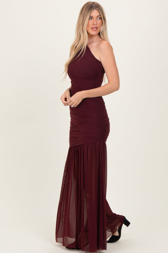 woman wearing a burgundy one-shoulder maxi dress with ruching and a sheer skirt, standing in profile