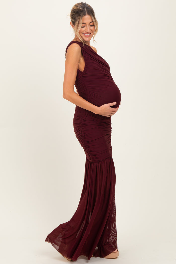 woman wearing a burgundy maternity gown with ruched detailing and a flowing skirt, holding her baby bump