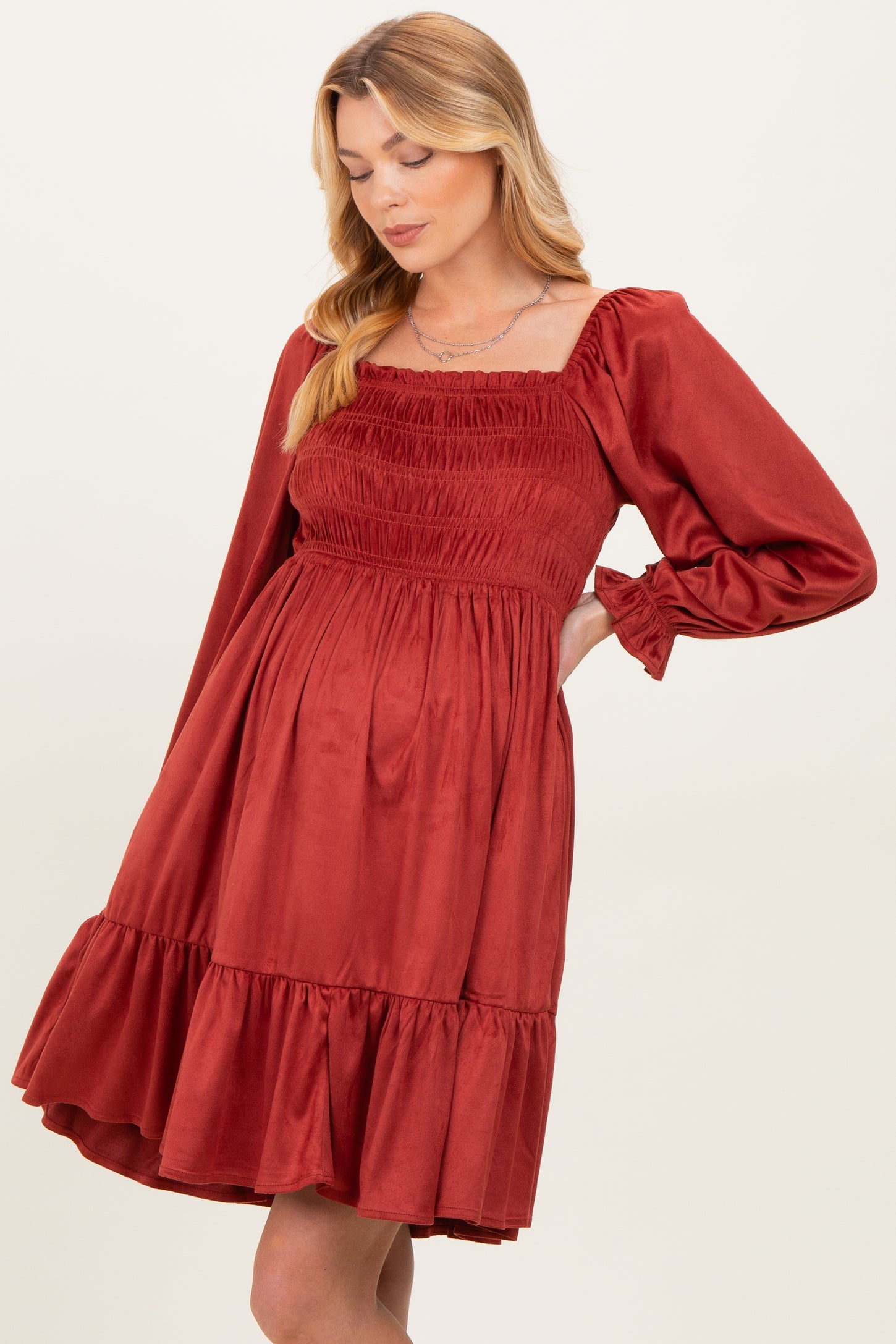 woman wearing a rust red maternity dress with smocked bodice and ruffled hem, gently showcasing her baby bump