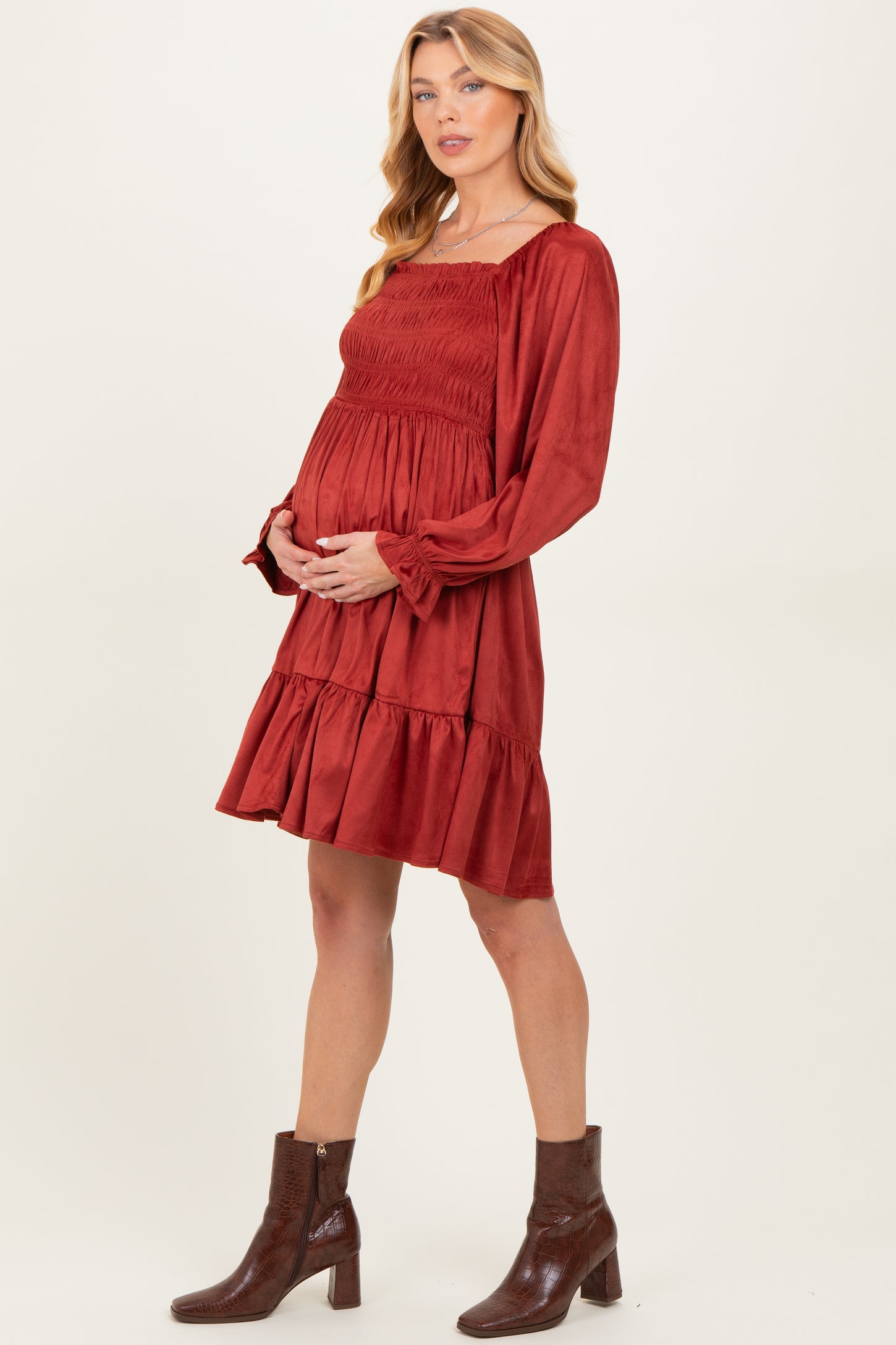 woman wearing a rust colored maternity dress with long sleeves, smocked bodice, and tiered skirt, holding her baby bump