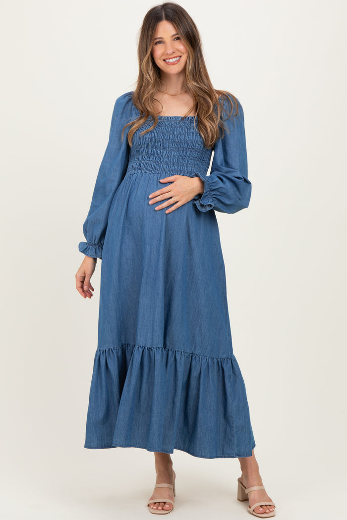 woman wearing a blue denim maternity dress with long sleeves and a tiered skirt, gently cradling her baby bump