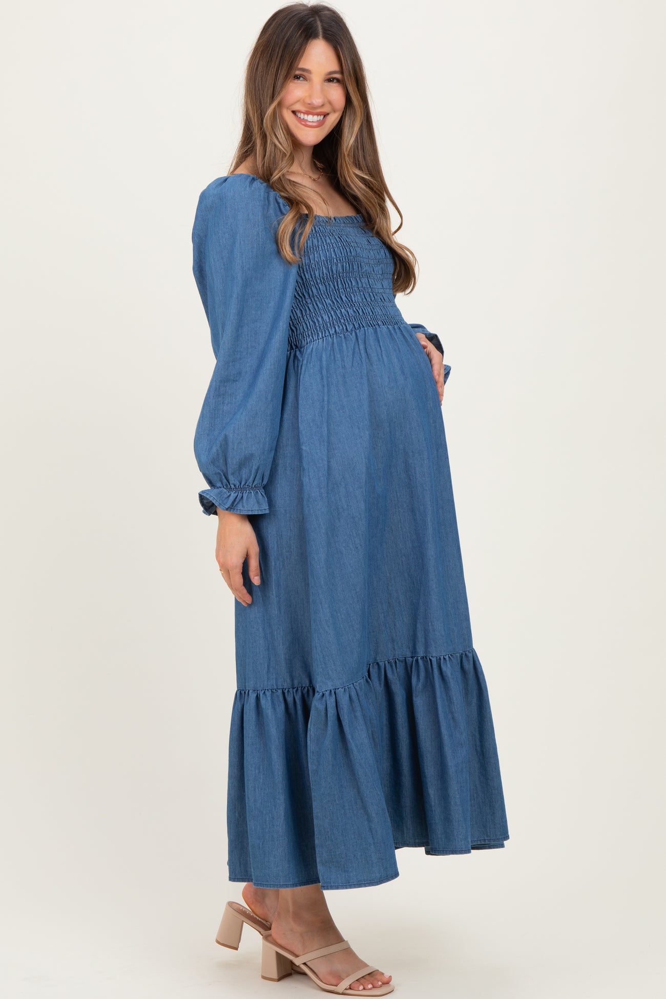 woman wearing a denim blue maternity dress with a smocked bodice and ruffled hem, holding her baby bump