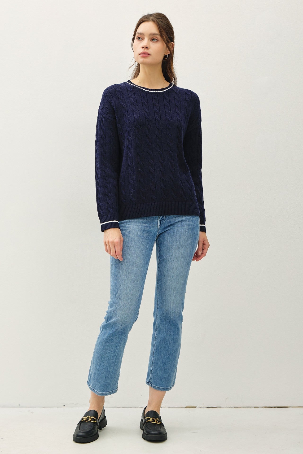 woman wearing a navy cable knit sweater with white trim and light blue cropped jeans, standing in a relaxed pose