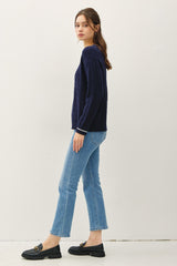 woman wearing a navy cable knit sweater, light blue cropped jeans, and black loafers, standing sideways against a white background