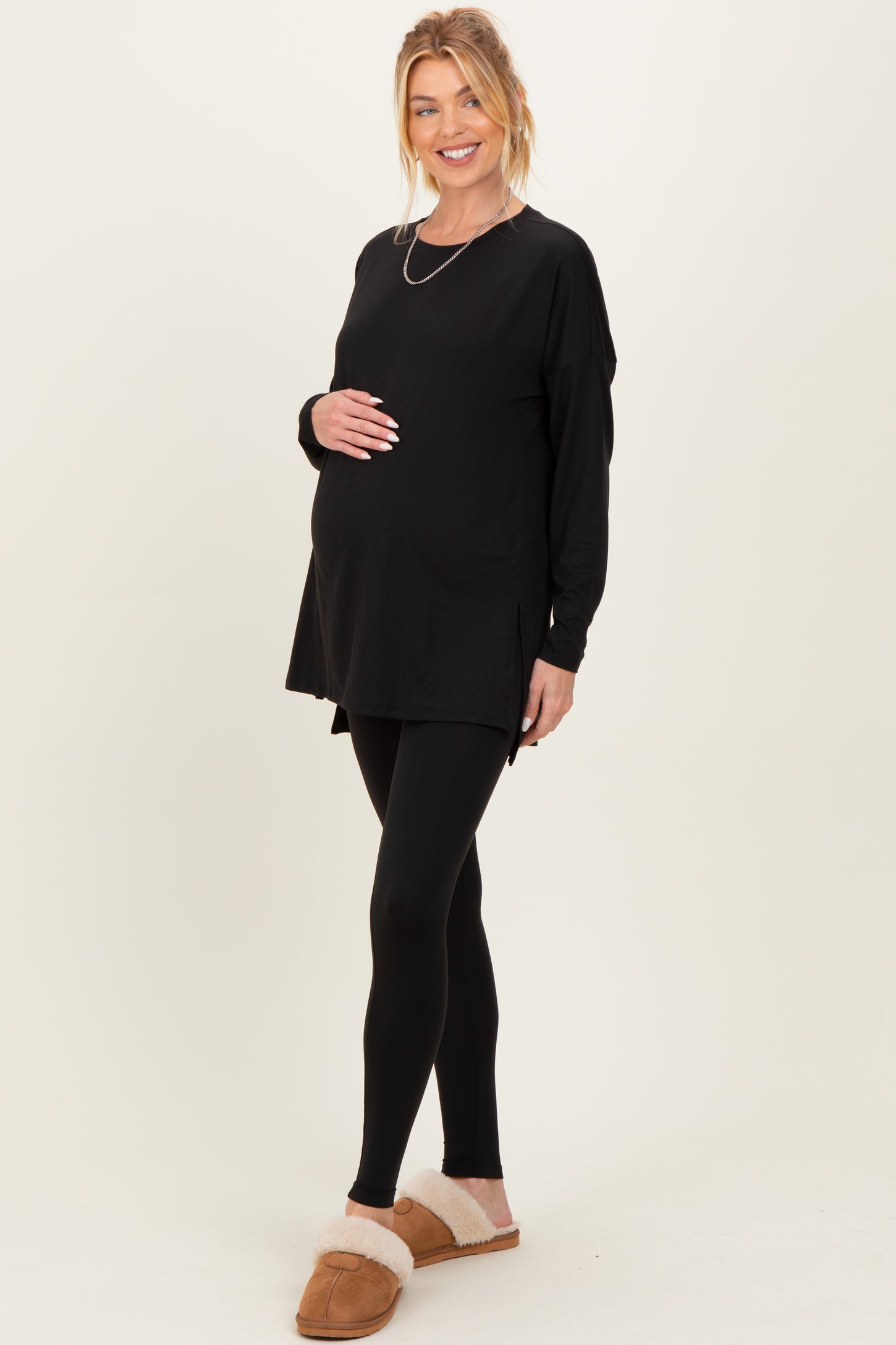 woman wearing a black long sleeve maternity top and black leggings, holding her baby bump with a smile