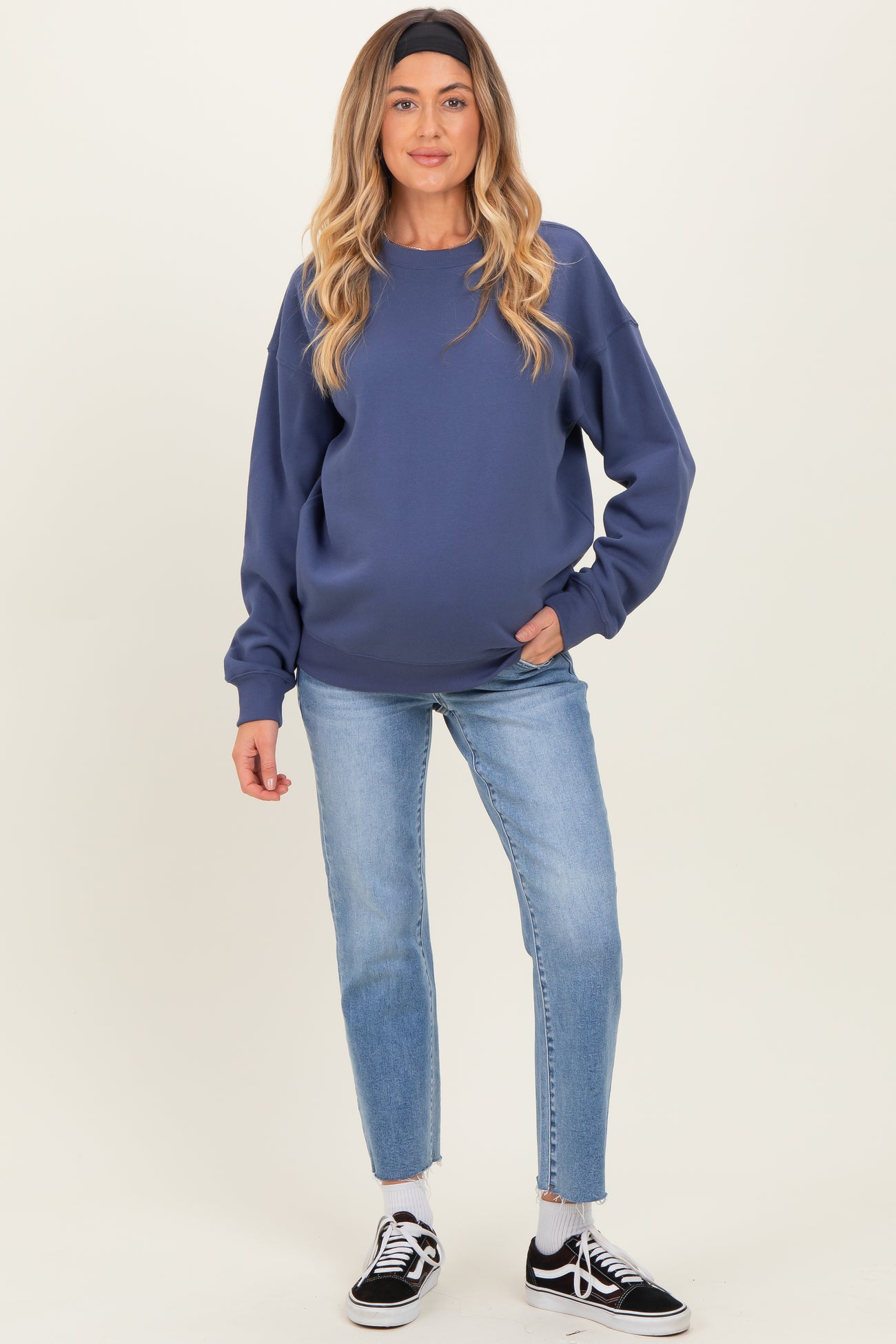 woman wearing a dark blue maternity sweatshirt and blue jeans, showing her baby bump, standing stylishly with a headband